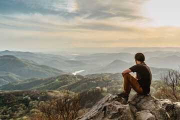 Naklejka na ściany i meble Male traveler with peaceful mind sitting on rock enjoying views of spring lush valley at sunset.Hiking day active lifestyle.Wanderlust woman relaxing outdoors.Travel freedom meditation concept