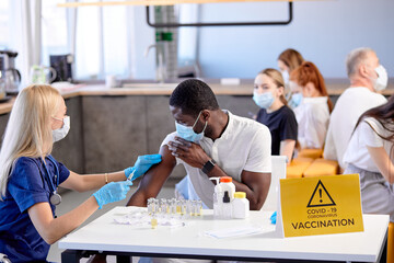 Obraz premium Mass vaccination of mixed race people in hospital office. nurse protective gear at work, puts a prick in african man's forearm, mass immunization, side view. copy space