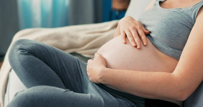 Close-up on profile of sitting pregnant women, living room scenery, girl dreamily touches her belly gently, strokes it, caring future mother, advanced pregnancy, motherly love
