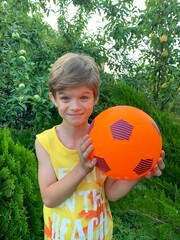 Funny smiling boy in a yellow T-shirt with a bright orange soccer ball on a background of green fruit trees. Children's ball games in nature in the fresh air.