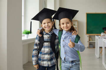 Elementary school learning and kids concept. Portrait of cheerful little student classmate wearing graduation cap standing in classroom and hugging smiling to camera showing thumbs up