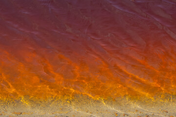 Textured surface of red poisoned muddy lake water near sandy shore in shallow water. There are small ripples on the water, there are reflections of light. Water pollution, background, copy space.