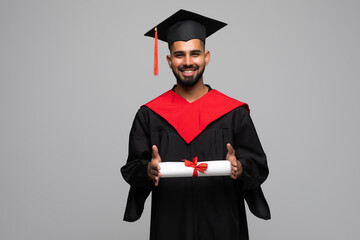 Smiling indian male graduate student in mortar board and bachelor gown with diploma over grey...