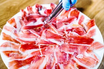 Ham plate. Appetizing traditional Spanish delicious slice Iberian jamón or cured ham leg. Person holding tweezers. Spanish gourmet food. Typical Spanish or Catalan food. Iberian pork. - obrazy, fototapety, plakaty