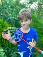 A cute cheerful boy holds a badminton racket in his hand. The child is going to play outdoors. Children's games in the summer camp.