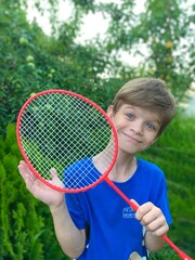 A funny boy with a badminton racket in his hands stands against the background of green fruit trees Children playing in the camp. The child rejoices in summer.