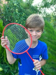 A little cute boy in a blue T-shirt holds a badminton racket in his hands. Child in the garden. Children's games in nature.
