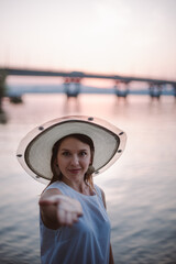 Fototapeta premium a young, happy woman in a straw hat stretches out her hand with an open palm to the camera and invites you to go on a summer vacation or travel to the river bank with a bridge at sunset. 