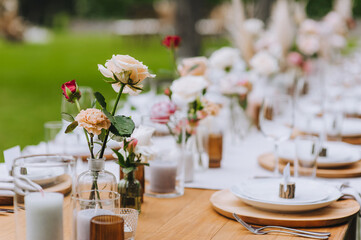 Beautiful multi-colored roses stand in a glass vase, a candle in a candlestick on the background of a wooden table with dishes at a banquet. Photography, concept.