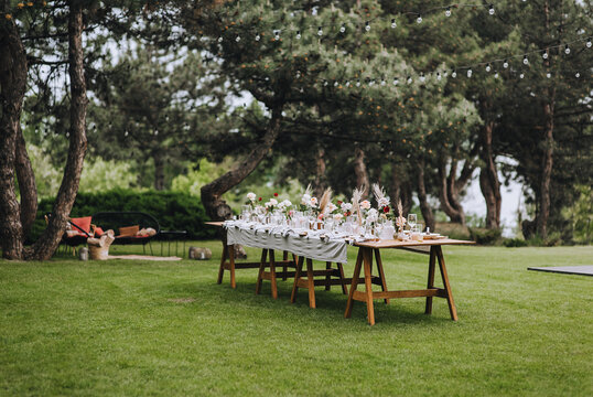 Wooden, long decorated with flowers, reeds, empty dishes, plates, forks, knives, glasses with a white tablecloth stands on the green grass in the park, forest. Wedding banquet.