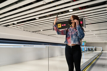 latina tourist woman with mask moving along the airport treadmill with social distancing while taking a selfie with her smart phone during the pandemic of coronavirus or covid19 virus