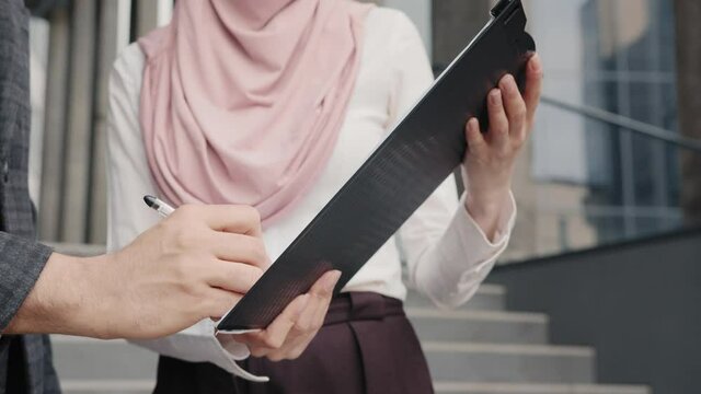 Woman in hijab holding clipboard while man signing contract. Arab man signing papers on clipboard that holding woman. Business colleagues in formal clothes standing on stairs outdoors near the office