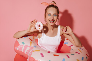 Obraz na płótnie Canvas Cheerful woman with round earrings and cool makeup in light swimsuit laughing and posing with donut and large swimming rings on pink backdrop..