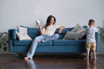 tired young mother or nanny sits on the couch and watches her son playing.