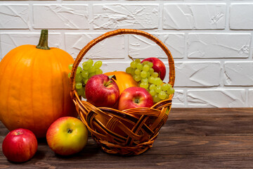 Orange pumpkins with apples and pears in a wicker basket on a brown wooden table against the background of a white brick wall.