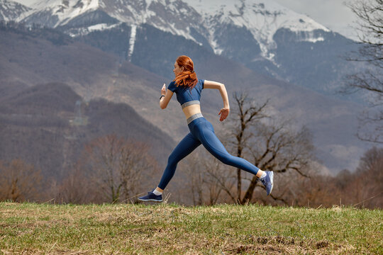 Side View Portrait Of Fit Redhead Lady Jumping High During Training In Nature, Sportswoman In Blue Sportive Outfit Enjoy Jogging, Mountains Landscape In The Background