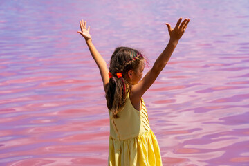 Happy little girl in yellow dress standing in water of pink salt lake on sunny summer day. Wanderlust travel concept