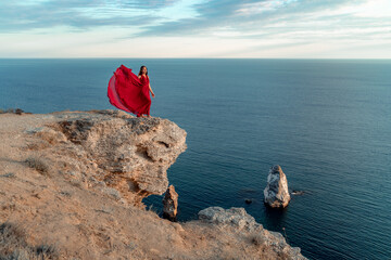 A girl with loose hair in a red dress stands on a rock rock above the sea. In the background, the sea and the rocks. The concept of travel