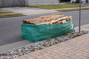 Green bag type dumpster filled with wood debris on a street near a sidewalk.