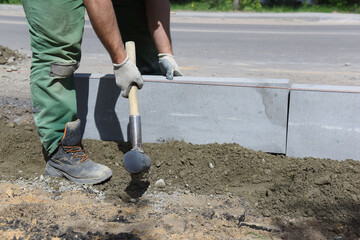 A worker knocks with a hammer on a concrete block, aligns curbs installed in cement, construction of a ring road.