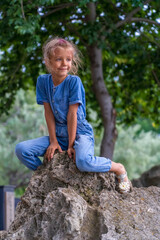 Naklejka na ściany i meble Little girl sitting on a huge stone in park. Cheerful childhood in nature.