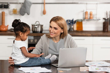smiling mother looking at adopted african american girl on table near gadgets