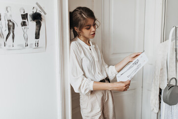 Woman dressed in white outfit looks at clothing samples. Dark-haired lady in light blouse and pants poses in cozy fashion designer office