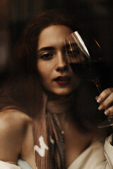 Attractive woman holds wine glass. Charming young girl in beige stylish dress looks into camera and poses in dark room
