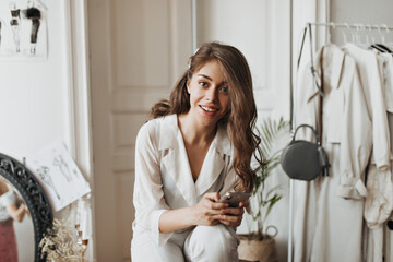 Woman with wavy hair looking into camera and holding phone. Brunette curly lady in white blouse...