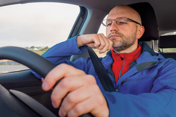 Tourist male driver in a blue jacket driving a car. The model is in his 40s, bald, grey short beard, wearing glasses.