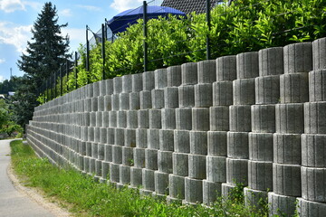 Stützmauer aus grauen runden Pflanzsteinen zur Hangsicherung und Abgrenzung eines Wohngrundstücks gegen die tiefer liegende Straße - obrazy, fototapety, plakaty