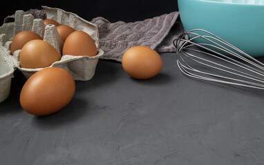 Fresh eggs in cardboard egg tray with whisk, plate and towel on dark background