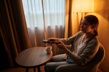 Evening rest at home. A young woman in casual clothes is sitting on an armchair. She watches TV....