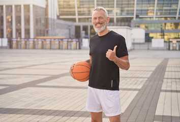 Active middle aged man in sportswear looking at camera, showing thumbs up, standing outdoors with...