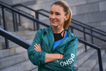 Portrait of attractive mature woman in sportswear smiling aside while posing outdoors after training in the city