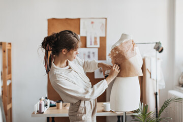 Lady in beige outfit designes clothes patterns. Brunette woman in light shirt creates stylish fashionable top on mannequin