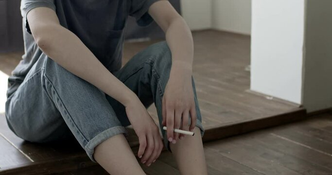 Close-up photo of female hands, lonely sad sitting smoking on floor, problem stress. Unhappy cropped woman in casual outfit sit with cigarette in hands, copy space. Desperate and depressed, at home