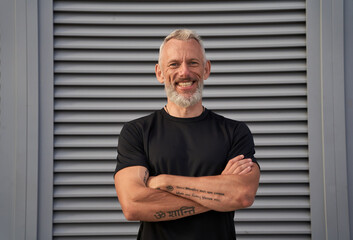 Portrait of mature man in perfect shape smiling at camera while standing with arms crossed outdoors...