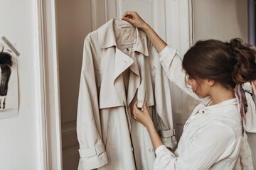 Lady looking at beige trench coat. Dark-haired woman in white blouse poses in white room and holds...