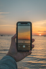 Hand holding a mobile phone taking a photo of the beautiful sunset on the beach