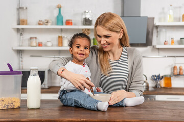 happy adopted african american kid holding spoon near bowl while sitting on kitchen table near smiling mother