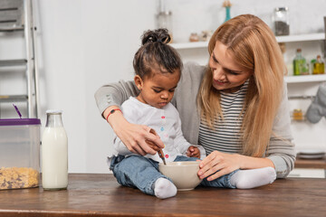 Fototapeta premium adopted african american kid holding spoon near bowl while sitting on kitchen table near smiling mother