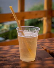 A plastic cup of apple cider with a biodegradable straw, standing on a wooden table. 