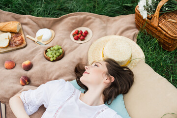 Top view of young woman lying near food and sun hat on blanket on grass