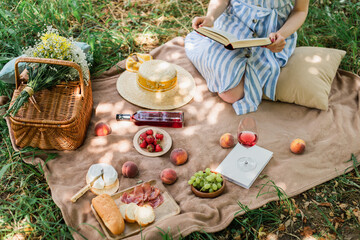 Cropped view of young woman holding book during picnic with wine and food in park - obrazy, fototapety, plakaty