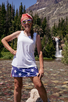 Fit woman wearing America USA clothing, poses at Running Eagle Falls waterfall in Glacier National Park