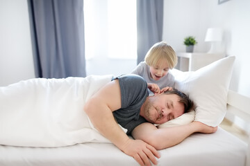 Little child awake, early in the morning he wakes up his sleeping father in bed at home. The tired...