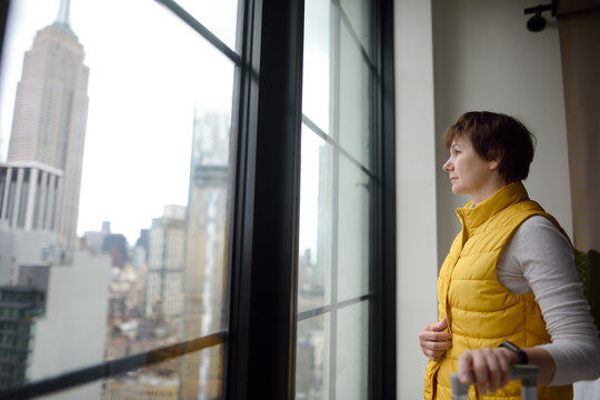 Woman tourist stays in hotel room in New York. Traveler admires of view the Empire State Building and skyscrapers of Manhattan outside the window. Booking apartment