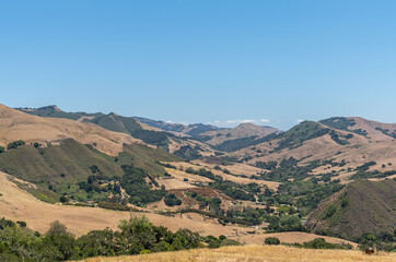 Fototapeta na wymiar Cambria, CA, USA - June 9, 2021: Wide landscape of Back country with dry ranch land and patches of green trees on steep flanks of mountains under blue sky.