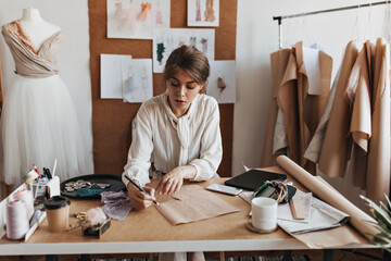 Portrait of lady drawing sketch and designing dress. Dark-haired woman in beige shirt creating...